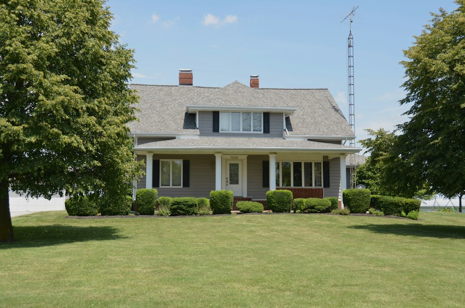 Two-story gray house with a full-width covered front porch, set back behind a wide green lawn and tall trees