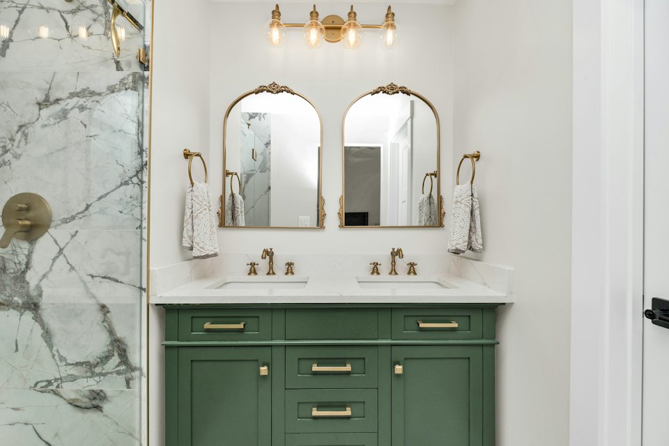 Bathroom with a green double vanity, two arched mirrors, brass fixtures and a marble-walled shower